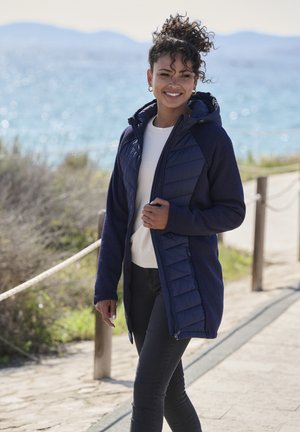 Marineblauer Kapuzenmantel aus strukturiertem Sweatmaterial; hat ein gestepptes Vorderteil und seitliche Reißverschlüsse. Getragen über einem cremefarbenen Pullover und schwarzen Hosen.