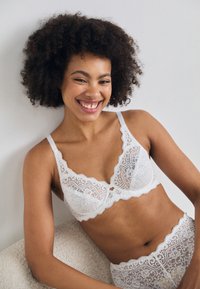 Il bralette bianco in pizzo e la biancheria coordinata presentano bordi scallop e motivi floreali, con una texture delicata e un design semitrasparente.