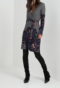 Langärmeliges Wickelkleid aus gemustertem Stoff in dunklen Farbtönen. Mit Taillengürtel und kontrastierenden geometrischen Mustern. Kombiniert mit schwarzen kniehohen Stiefeln.