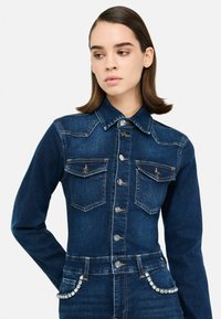 Giacca in denim scuro con una silhouette aderente, colletto classico, chiusura frontale con bottoni, due tasche sul petto e dettagli decorativi in strass in vita.