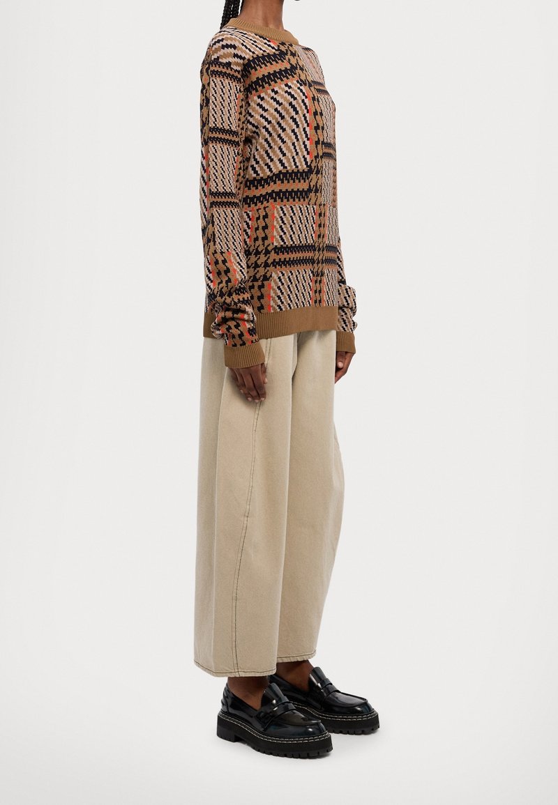 Personne portant un pull à motifs marron et noir, un pantalon large beige, et des mocassins noirs épais, debout de profil devant un fond uni.
