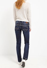 Jeans de mezclilla de un azul oscuro con un corte ajustado, que presentan un lavado sutil, costuras decorativas en los bolsillos traseros, y combinados con una camiseta deportiva ligera y zapatos.