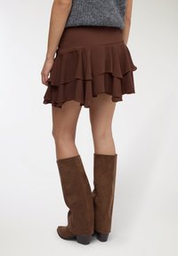 Gonna marrone a balze in tessuto leggero, con vita aderente. Abbinata a stivali marroni in suede alti fino al ginocchio, con texture liscia e ruvida.
