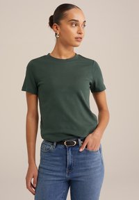 Zeleni kratkosleppi majica iz mešanice bombaža. Okrogla izreza, oprijet kroj, v kombinaciji z modrimi kavbojkami in črnim pasom z zlatim sponko.