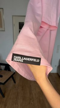 Hånd som holder et rosa Karl Lagerfeld Jeans-plagg med svart-hvitt merkebelagt kant på stoffet innendørs.