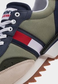 Sneaker che presenta una combinazione di tela verde e camoscio beige, con accenti blu navy e un dettaglio a strisce rosse, bianche e blu navy sul lato.