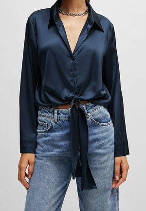 Žena v tmavě modré saténové košili s dlouhým rukávem uvázané v pase, světle modrých džínách a stříbrném řetízkovém náhrdelníku.