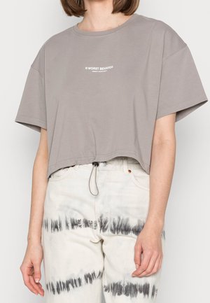 Osoba ubrana w luźny, szary, krótki t-shirt z napisem „WORST BEHAVIOR” oraz białe spodnie z czarnymi, poziomymi paskami typu tie-dye.