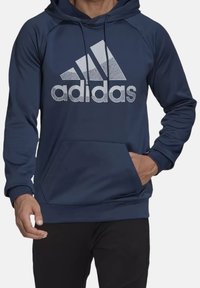 Sweat à capuche bleu marine avec poche avant, arborant un grand logo Adidas gris clair. Fabriqué en matériau doux et léger avec des poignets côtelés.