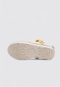 Suela de zapatilla beige con patrón de huella geométrica y cordón amarillo, mostrada contra un fondo gris claro liso.