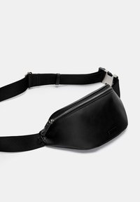Crni fau kožni bum bag sa zakrivljenim dizajnom, zatvaranjem na patentni zatvarač, podesivim crnim remenom i detaljima od srebrne boje.
