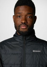 Giacca nera isolata con colletto alto e zip frontale. Presenta il logo Columbia nella zona sinistra del petto. Texture liscia e lucida.