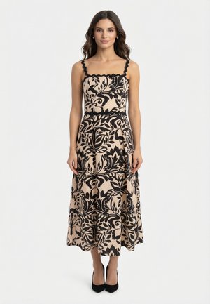 Femme portant une robe midi beige à motifs floraux noirs, avec des bretelles festonnées et des talons hauts noirs, debout sur un fond blanc.