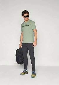 Zeleno DYNAFIT majica, črne športne hlače, refleksna sončna očala, črn nahrbtnik in večbarvni tekaški čevlji. Minimalistična zasnova, funkcionalna oblačila.