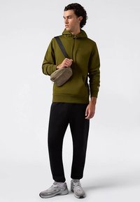 Olivovo zelená mikina s kapucňou a šnúrkami, čierne tepláky a sivé tenisky. Modelka nesie sivú kabelku cez telo.