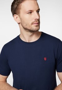 Granatowa bawełniana koszulka z okrągłym dekoltem i małym czerwonym logo na lewej piersi. Gładka faktura, dopasowany krój, krótkie rękawy.