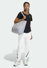 Bolso tote gris con una textura suave y diseño minimalista. Incluye correas resistentes y es sostenido por una persona que lleva una camiseta negra y pantalones blancos.