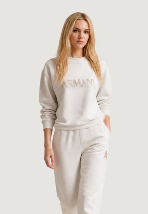 Femme blonde portant un sweat-shirt Armani beige clair et un pantalon de survêtement assorti, debout avec une main dans la poche devant un fond uni.