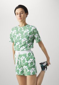 Conjunto de camisa blanca y pantalones cortos con patrón floral verde; material de algodón, ajuste holgado, cinturilla elástica y cordón. Zapatillas negras y azul verdoso.