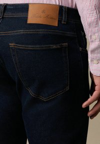 Jeans in denim scuro con una toppa in pelle marrone sulla vita posteriore, cuciture dorate e tasche posteriori standard.