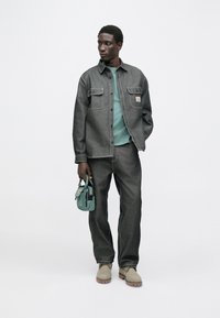 Camisa de mezclilla gris con dos bolsillos en el pecho, sudadera verde debajo, pantalones de mezclilla gris y zapatos de tono beige claro. Sosteniendo un pequeño bolso color teal.