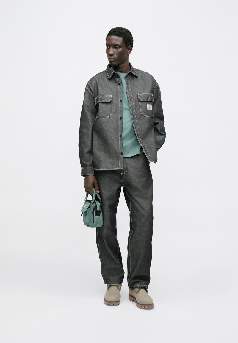 Camicia di denim grigio con due tasche sul petto, felpa verde sotto, pantaloni di denim grigio e scarpe color beige chiaro. Tiene in mano una piccola borsa turchese.