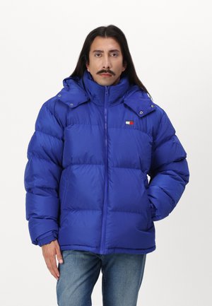 Chaqueta acolchada de color azul royal con un alto cuello, cierre de cremallera y capucha desmontable. Presenta un pequeño parche con el logo en el pecho y puños elásticos.