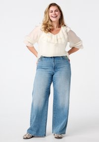 Witte blouse met ruches en doorschijnende mouwen, gecombineerd met hoge, wijdvallende blauwe jeans. De schoenen hebben een gestructureerd, patroonontwerp.