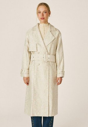 Mujer con abrigo largo beige con estampado, cinturón ancho, hebilla y grandes solapas sobre un top crema de cuello alto y vaqueros azul oscuro.