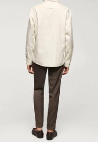 Persona che indossa una camicia a maniche lunghe color crema con bottoni, pantaloni marroni a sigaretta con orli e scarpe eleganti marroni scure, in piedi contro uno sfondo neutro.