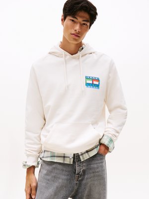 Felpa bianca con tasca frontale, adornata da un logo stampato colorato. Indossata sopra una camicia a quadri verde e abbinata a jeans grigi.