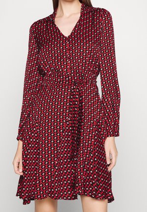 Frau trägt ein rot-schwarz gemustertes Kleid mit langen Ärmeln, taillierter Taille und einer Knopfleiste vorne.