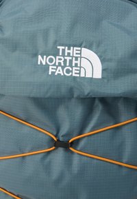 Γκρι υφασμάτινο σακίδιο πλάτης με το λογότυπο The North Face, με ρυθμιζόμενο κορδόνι bungee σε πορτοκαλί και υφή πλέγματος στο υλικό.