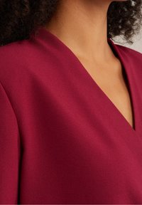 Vestito bordeaux con scollatura a V profonda, realizzato in tessuto liscio e strutturato; presenta maniche ampie e una leggera lucentezza.