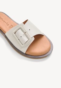 Sandale blanche en cuir à bride large avec une grande boucle carrée, semelle intérieure matelassée couleur fauve et semelle à bordure décorée sur fond blanc.