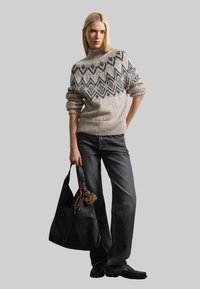 Maglione lavorato a maglia beige con motivo geometrico nero, abbinato a jeans scuri e una borsa in pelle nera con un accento di pom-pom in pelliccia.