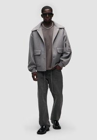 Šedá vlna smíšená bunda s přehnaným límcem a předními kapsami, vrstvená na béžové mikině, doplněná volnými šedými džíny a černými botami.