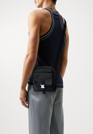 Black crossbody bag with textured fabric, zip closure, and adjustable strap, featuring a silver buckle and logo detail.