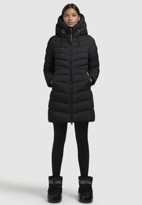 Zwarte pufferjas met een capuchon, verticaal gestikt patroon, ritssluiting aan de voorkant, zijzakken en een middellange ontwerp. Gecombineerd met zwarte leggings en laarzen.