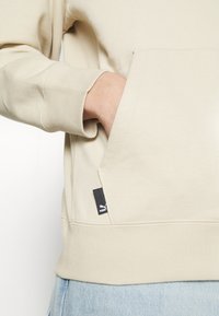 Main dans la poche avant d'un sweat à capuche beige, poignet côtelé, étiquette noire avec le logo Puma visible, jean bleu clair à l'ourlet.