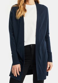 Marinblå stickad kofta med ribbad öppen front, långa ärmar och ribbade muddar. Bärs över en vit t-shirt och svarta byxor.