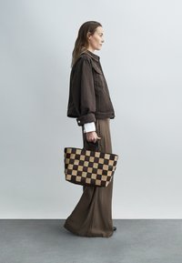Sac fourre-tout à damier marron et beige, de forme structurée, avec deux poignées et une texture tissée. Le modèle porte une veste et un pantalon marron.