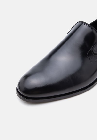 Μαύρο γυαλιστερό δερμάτινο παπούτσι φόρμας χωρίς κορδόνια, με στρογγυλεμένη μύτη και λείο πάνω μέρος σε απλό ανοιχτό φόντο.