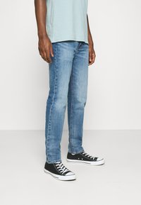 Jeans in denim azzurro chiaro con vestibilità dritta, caratterizzati da un lavaggio sbiadito e un design a cinque tasche, abbinati a sneakers nere basse.