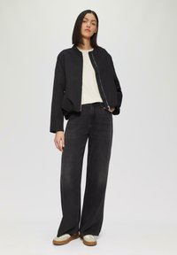 Veste bomber noire avec fermeture éclair, associée à un jean noir à jambes larges et des chaussures à lacets beiges. Design minimaliste, texture de tissu lisse.