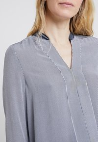 Žena je oblečena v dlouhý rukáv, tenkým pruhovaným tmavě modrým a bílým halenkou s výstřihem do V a malým stojacím límcem.