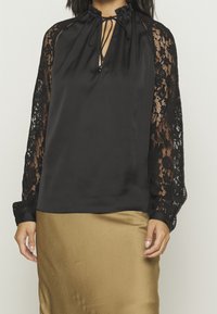 Blouse noire avec manches longues en dentelle, fermeture à nouer sur le devant et corps lisse en satin. Associée à une jupe en satin doré.