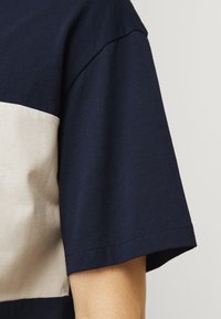 Mørkeblå T-shirt med en cremefarvet vandret stribe, lavet af bomuld. Korte ærmer, rund halsudskæring, synlige stikninger ved kanten.