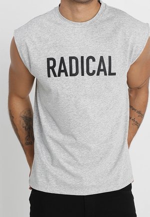 Mand iført ærmeløs grå skjorte med ordet "RADICAL" i fede sorte bogstaver på tværs af brystet og tatoveringer på begge arme.