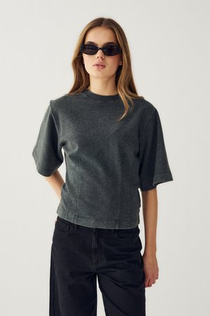 Žena s dlhými vlasmi, ktorá nosí čierne obdĺžnikové slnečné okuliare, tmavosivé tričko s krátkym rukávom a čierne nohavice, stojaca pred jednoduchým svetlým pozadím.